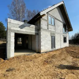 Surowy stan domu z białych bloczków betonowych, z widocznym zadaszonym miejscem na garaż, czarnym dachem i oknami, otoczony piaszczystym terenem budowy w słoneczny dzień.