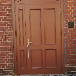 Brązowe, dwuskrzydłowe drzwi wejściowe z łukowym nadprożem z cegły, widoczne z zewnątrz, z metalową kratką przed progiem.