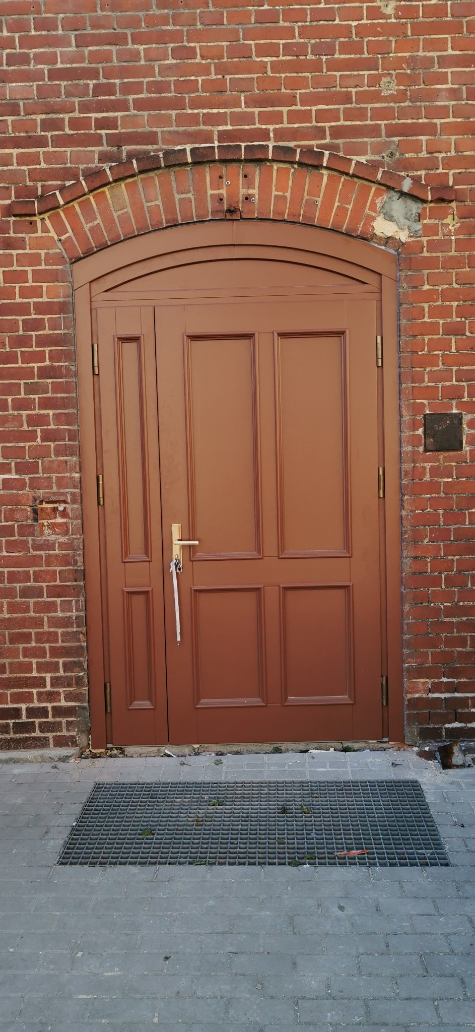 Brązowe, dwuskrzydłowe drzwi wejściowe z łukowym nadprożem z cegły, widoczne z zewnątrz, z metalową kratką przed progiem.