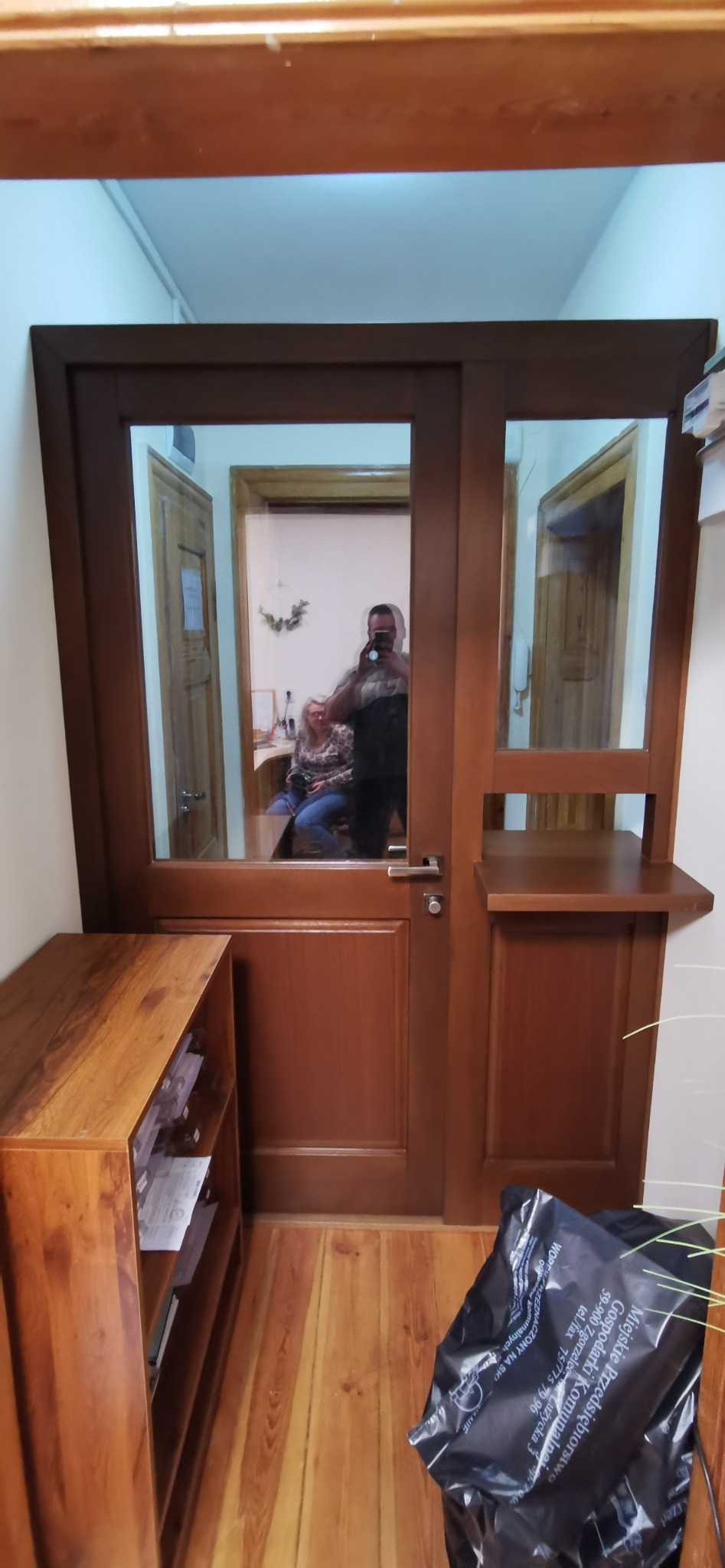 Brązowe drzwi wewnętrzne z szybami i półką, widoczne odbicie osoby robiącej zdjęcie oraz pomieszczenie biurowe w tle.