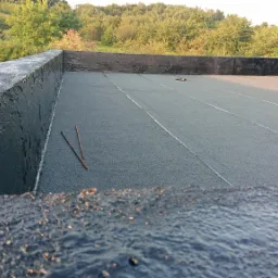 Płaski dach pokryty papą termozgrzewalną, z widocznymi detalami obróbki krawędzi i dwoma prętami leżącymi na powierzchni, na tle zieleni i fragmentu dachu z dachówką.