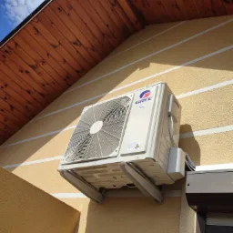 Klimatyzator marki Gree zamontowany na elewacji domu z drewnianą podbitką dachową, widoczne przewody i mocowania.