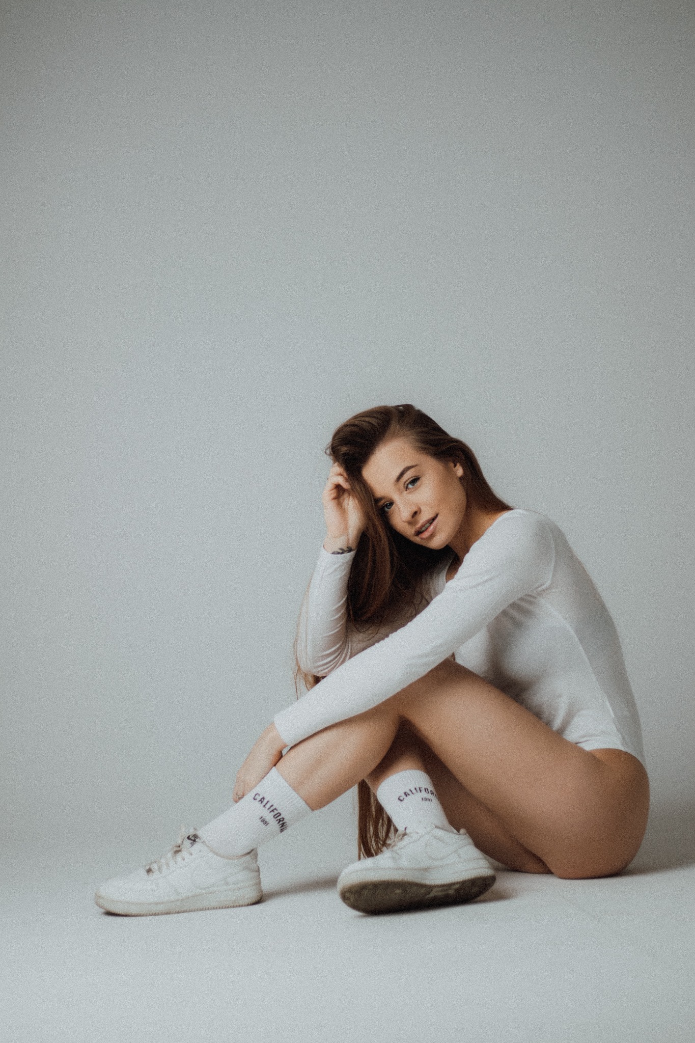 Portret młodej kobiety w białym body i skarpetkach California, siedzącej na podłodze na jasnym tle, z ręką we włosach i delikatnym uśmiechem.