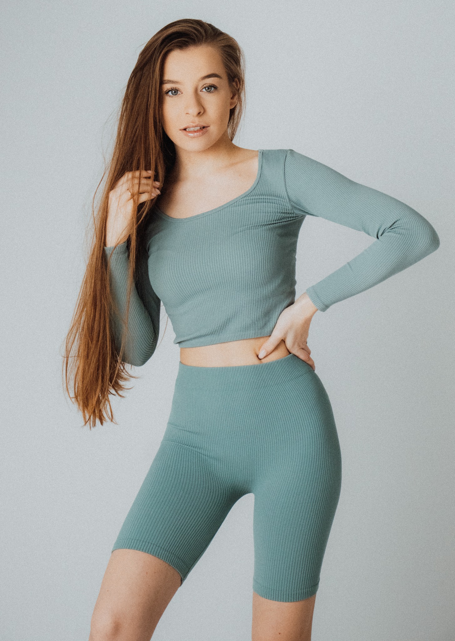 Portret kobiety w prążkowanym, turkusowym komplecie sportowym, z ręką na biodrze i długimi, rudymi włosami opadającymi na ramię, na jasnym tle.
