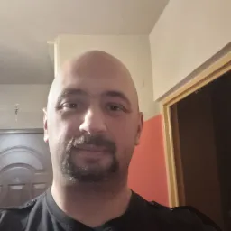 Portret mężczyzny z ogoloną głową i ciemną brodą, ubrany w czarną koszulkę, na tle ściany w odcieniach beżu i pomarańczy oraz brązowych drzwi.