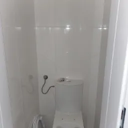 Biała toaleta z otwartą deską w małej, wykafelkowanej łazience z szarymi płytkami podłogowymi i widocznym wężem doprowadzającym wodę.