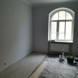 Pokój po malowaniu ścian na szaro, widoczne zabezpieczenia podłogi folią malarską, okno łukowe z grzejnikiem, listwy przypodłogowe do montażu i akcesoria malarskie.
