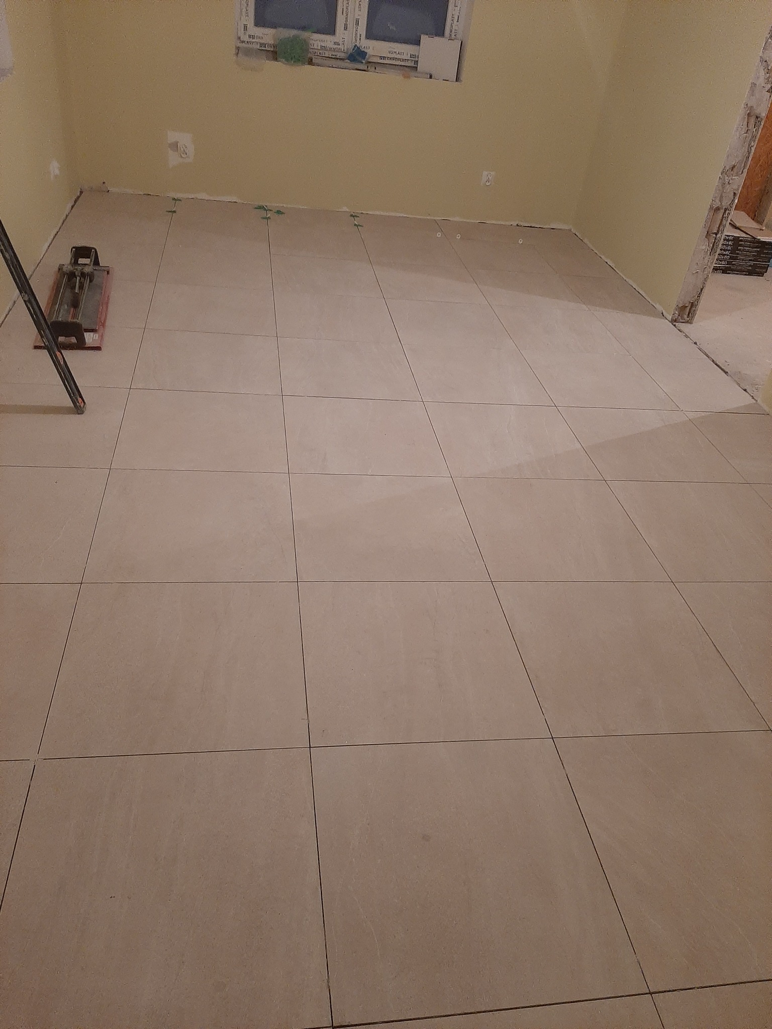 Ułożone beżowe płytki podłogowe w trakcie prac, z widoczną maszynką do cięcia płytek i elementami poziomowania, surowe ściany w tle.