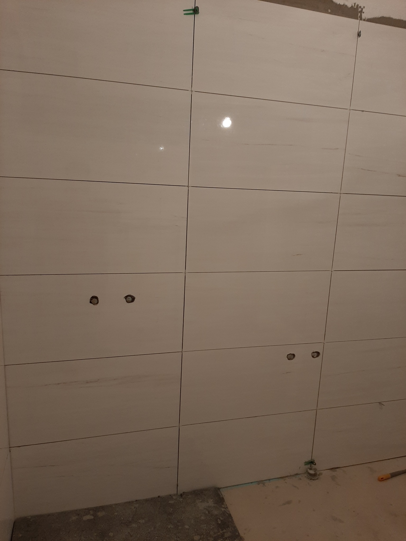 Ściana w trakcie układania jasnych, prostokątnych płytek ceramicznych imitujących marmur, z widocznymi otworami pod instalacje i zielonymi krzyżykami dystansowymi, częściowo wykończona z widocznymi...