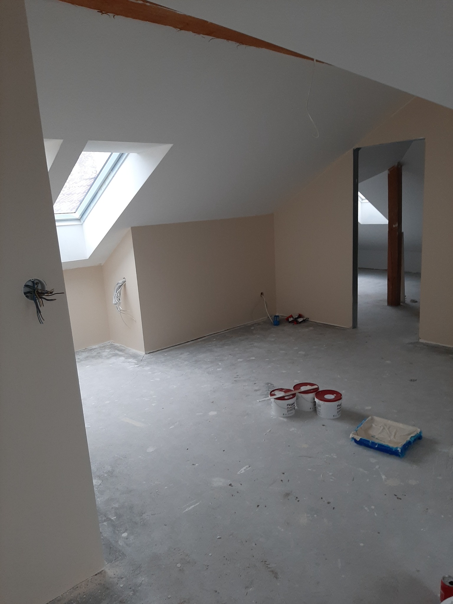 Poddasze w trakcie prac wykończeniowych: pomalowane ściany w odcieniach beżu, okno dachowe, puszki z farbą i kuweta malarska na szarej podłodze, widoczne kable elektryczne.
