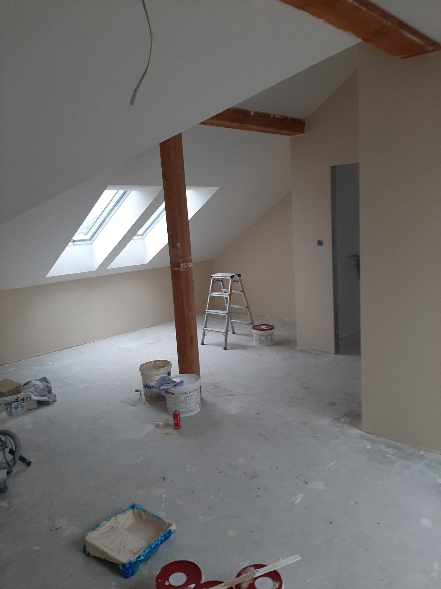 Poddasze po malowaniu ścian na jasny beż, widoczna drewniana belka, okna dachowe, drabina malarska, puszki z farbą i narzędzia malarskie na podłodze.
