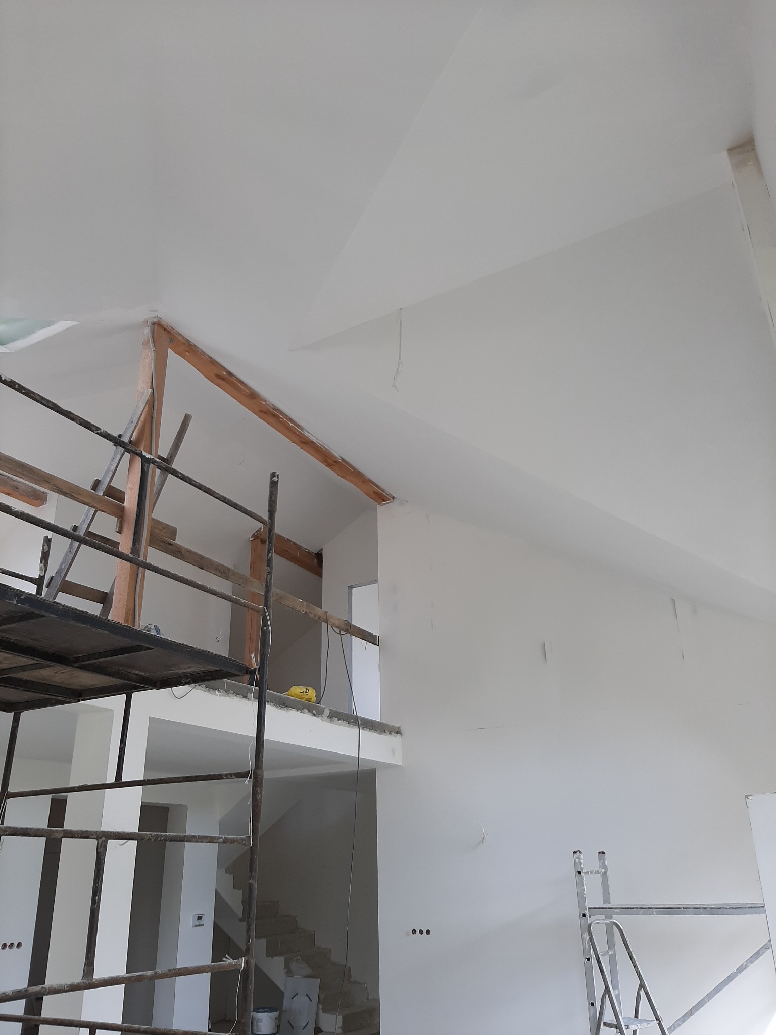 Wnętrze w trakcie malowania: białe ściany, drewniana konstrukcja, rusztowania, widoczne schody i otwory drzwiowe, sufit ze skosem.