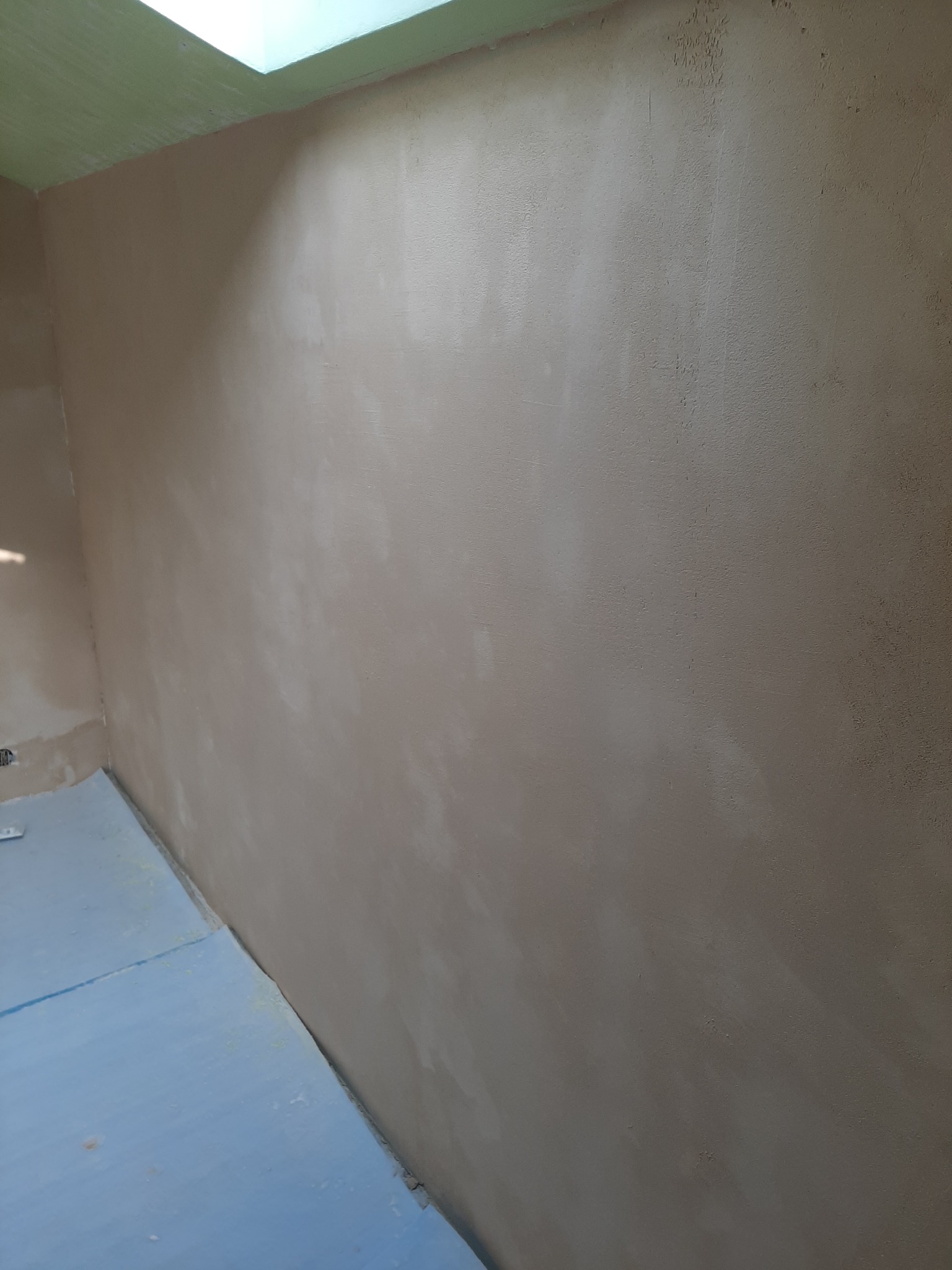 Ściana po nałożeniu gładzi, widoczne nierówności i faktura, przygotowana do dalszych prac wykończeniowych, z boku fragment niebieskiego zabezpieczenia podłogi.