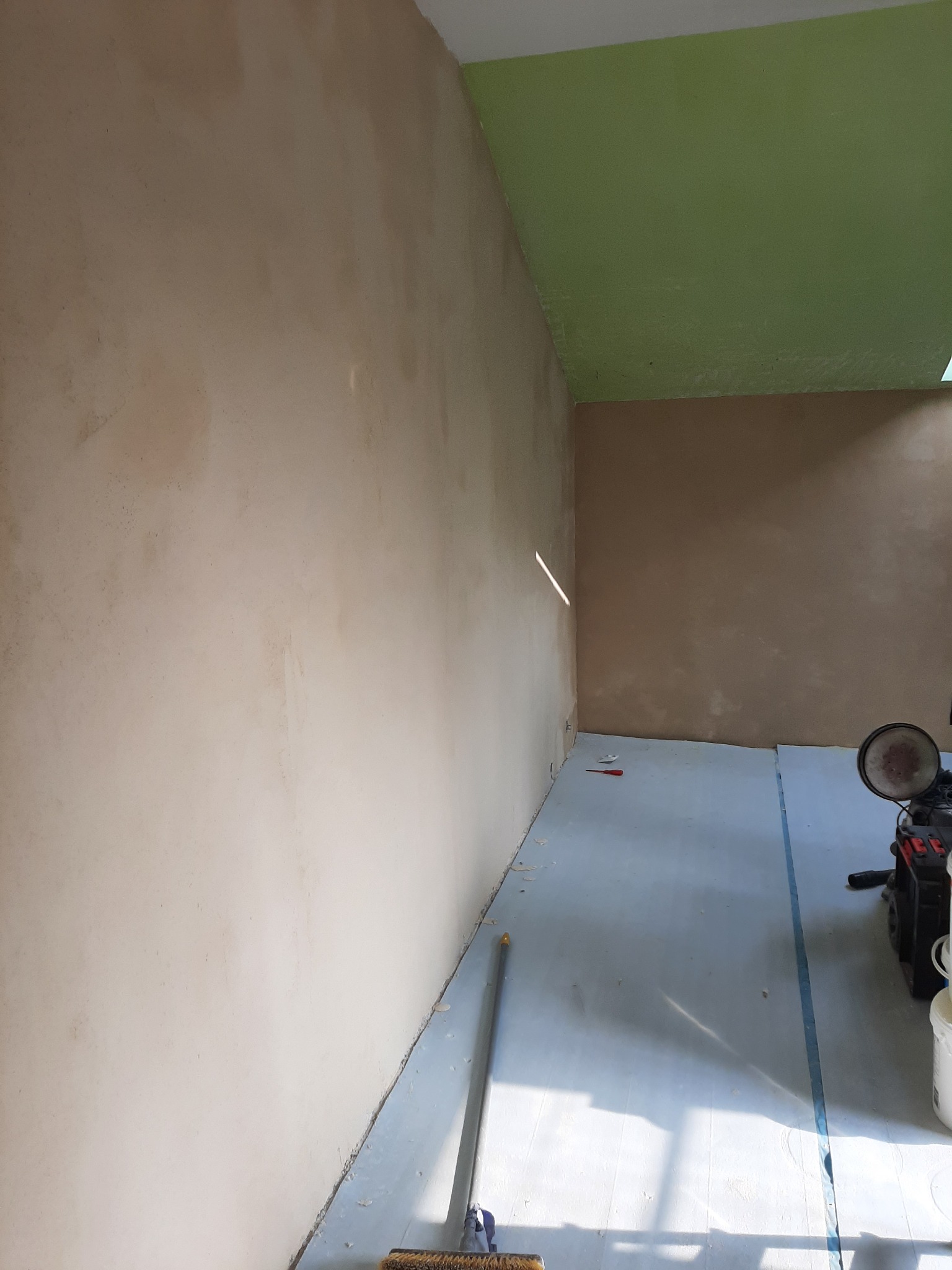 Ściana w trakcie gładzenia, widoczne nierówności i ślady po szpachlowaniu, podłoga zabezpieczona folią, w tle zielony sufit i sprzęt budowlany.