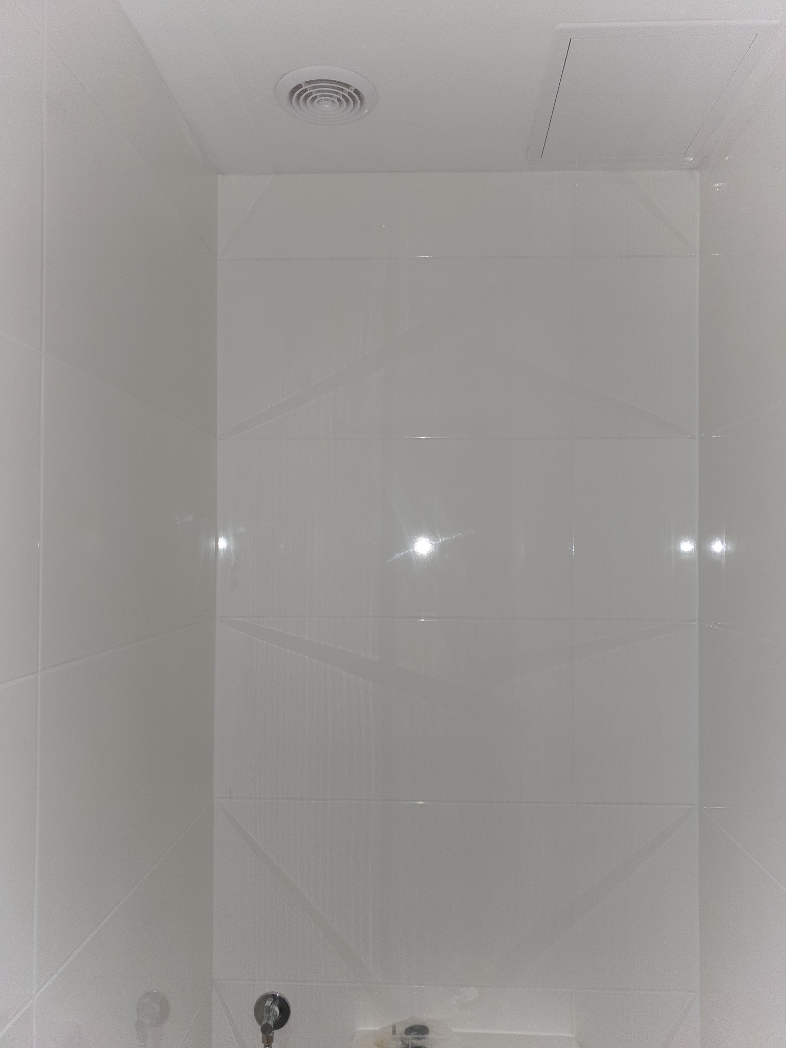 Białe, błyszczące płytki ceramiczne ułożone w łazience z widocznym otworem wentylacyjnym w suficie i klapą rewizyjną.