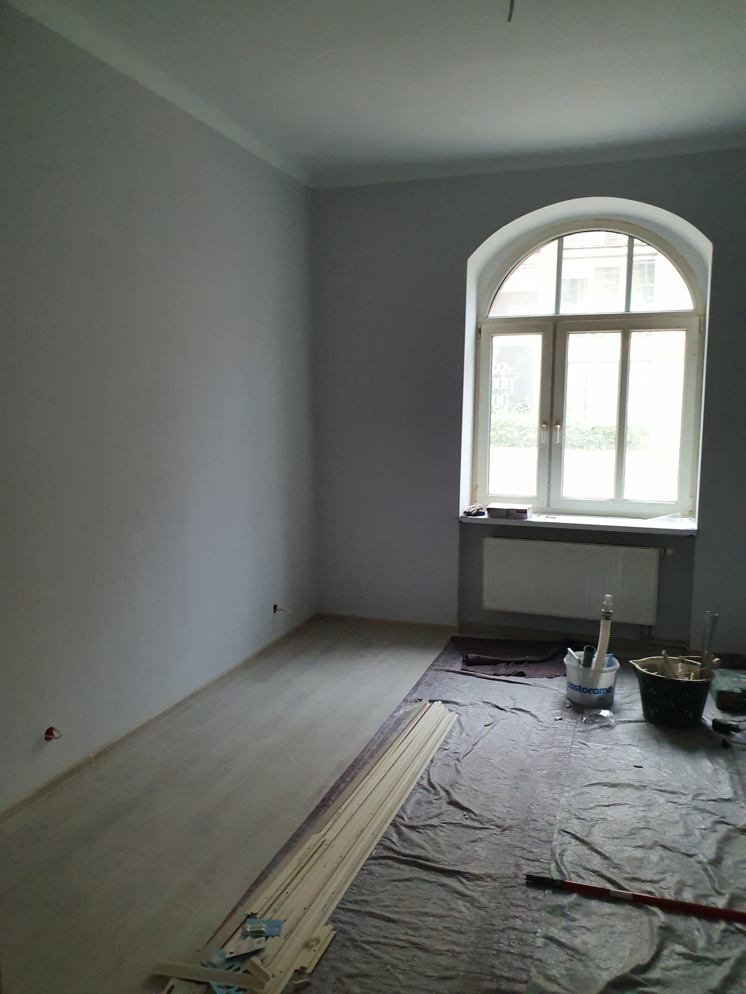 Pokój po malowaniu ścian na szaro, widoczne zabezpieczenia podłogi folią malarską, okno łukowe z grzejnikiem, listwy przypodłogowe do montażu i akcesoria malarskie.