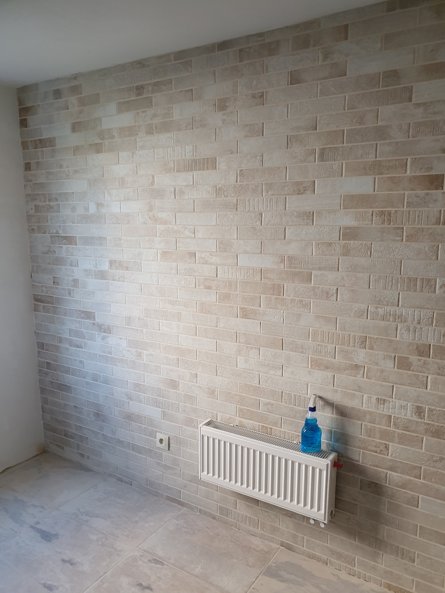 Ściana wyłożona beżową cegłą dekoracyjną z widocznym grzejnikiem i butelką z płynem do czyszczenia, gniazdko elektryczne na ścianie, podłoga wyłożona płytkami imitującymi kamień.