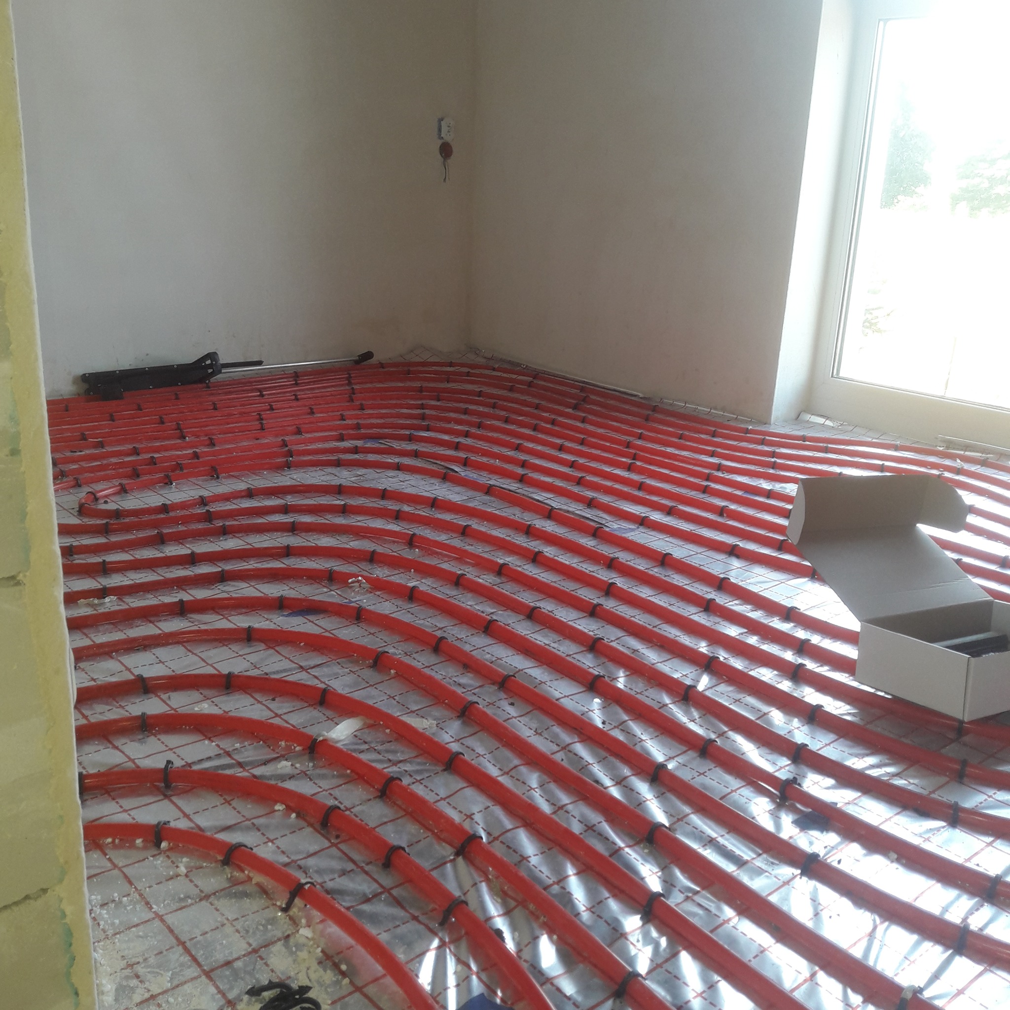 Czerwone rury ogrzewania podłogowego ułożone na srebrnej folii izolacyjnej w jasnym pomieszczeniu z oknem, widoczny karton z elementami montażowymi i narzędzie do mocowania rur.