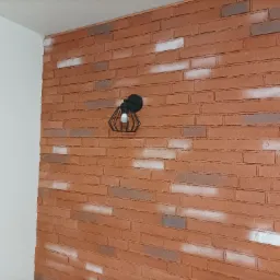 Dekoracyjna ściana z cegły klinkierowej w odcieniach czerwieni i brązu, miejscami pobielana, oświetlona czarnym, ażurowym kinkietem z żarówką Edison, sąsiadująca z białą ścianą.