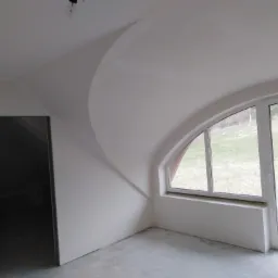 Wnętrze pokoju ze świeżo pomalowanymi ścianami na biało, z nietypowym łukowym przejściem sufitu i oknem z widokiem na zewnątrz, podłoga betonowa.