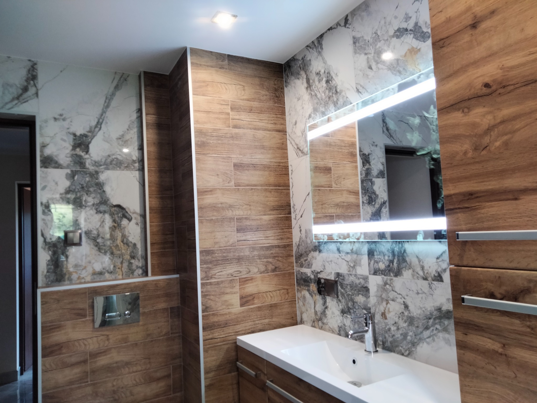 Nowoczesna łazienka z marmurowymi i drewnopodobnymi płytkami, podświetlanym lustrem nad białą umywalką z szafką, widoczny fragment toalety z chromowanym przyciskiem spłukiwania.