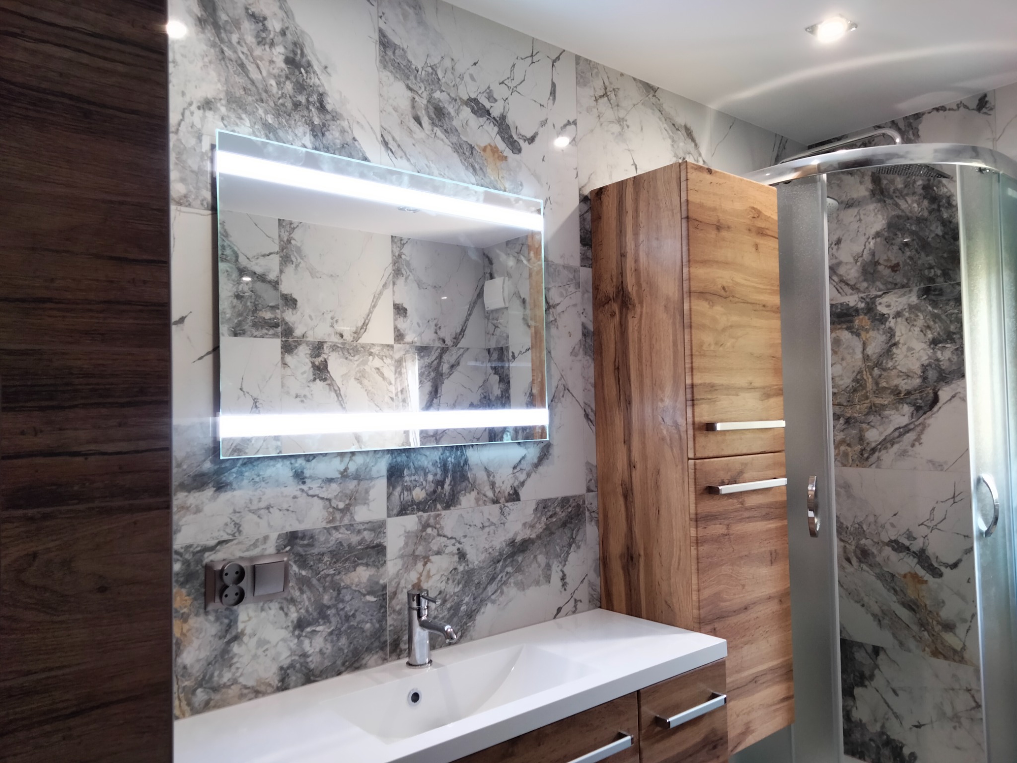 Nowoczesna łazienka z marmurowymi płytkami, podświetlanym lustrem, drewnianą szafką i kabiną prysznicową z chromowanymi detalami.