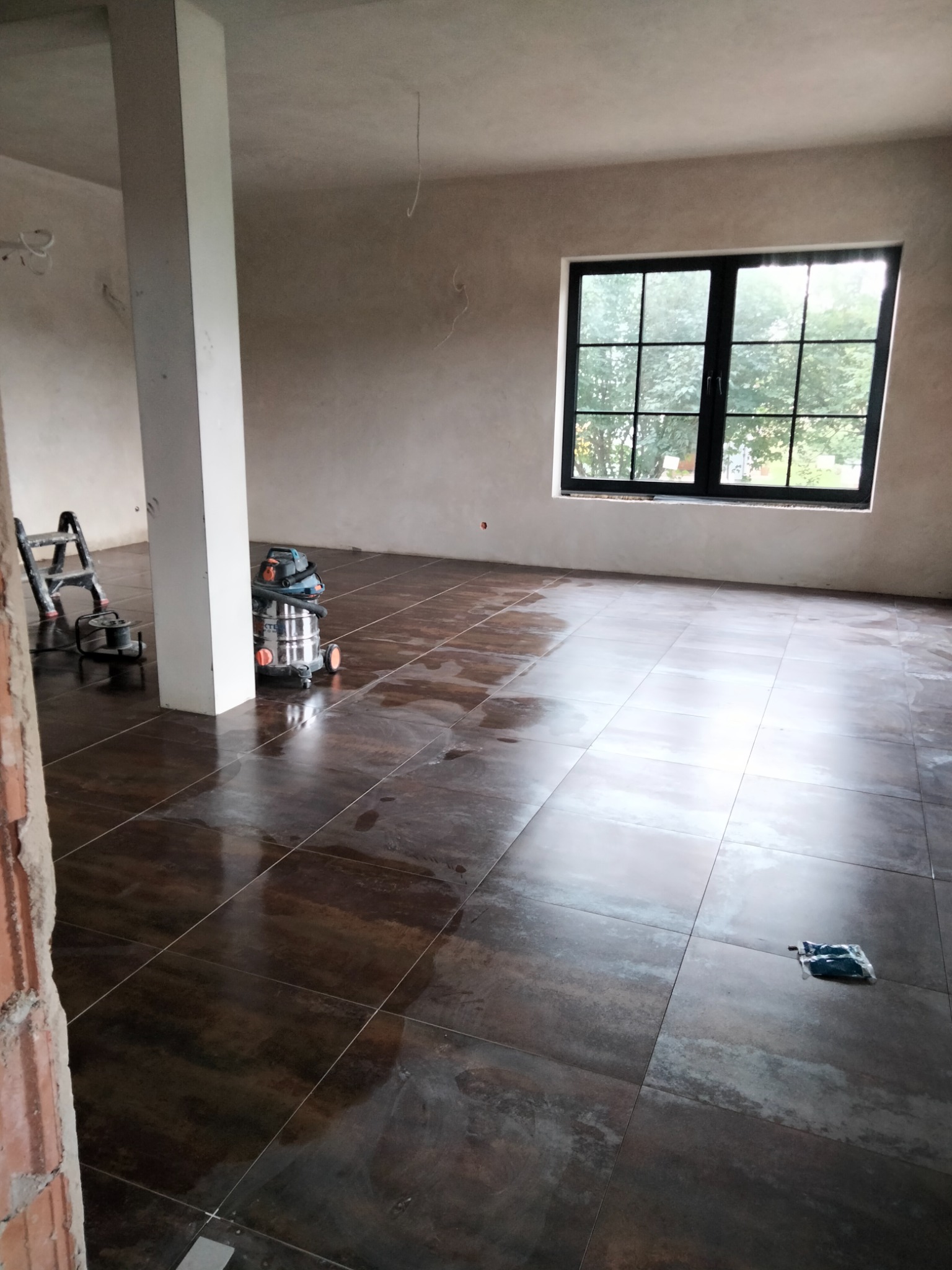 Układanie ciemnych, połyskliwych płytek podłogowych w Sanoku, widoczny odkurzacz przemysłowy i drabina, pomieszczenie w trakcie wykańczania.