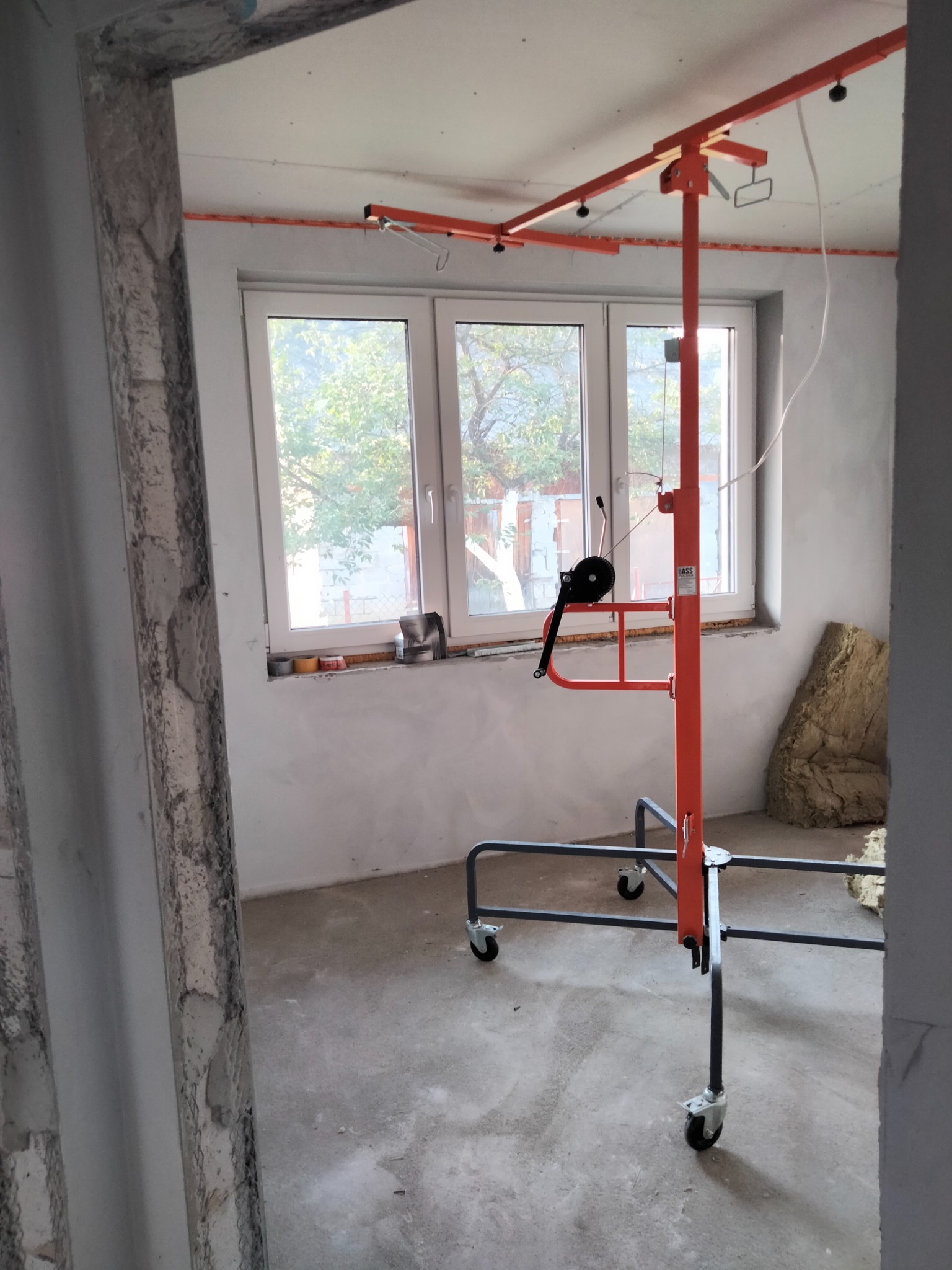 Pomarańczowy podnośnik do płyt gipsowo-kartonowych na kółkach, ustawiony w surowym wnętrzu z oknem, widoczne ślady po pracach wykończeniowych i materiał izolacyjny.