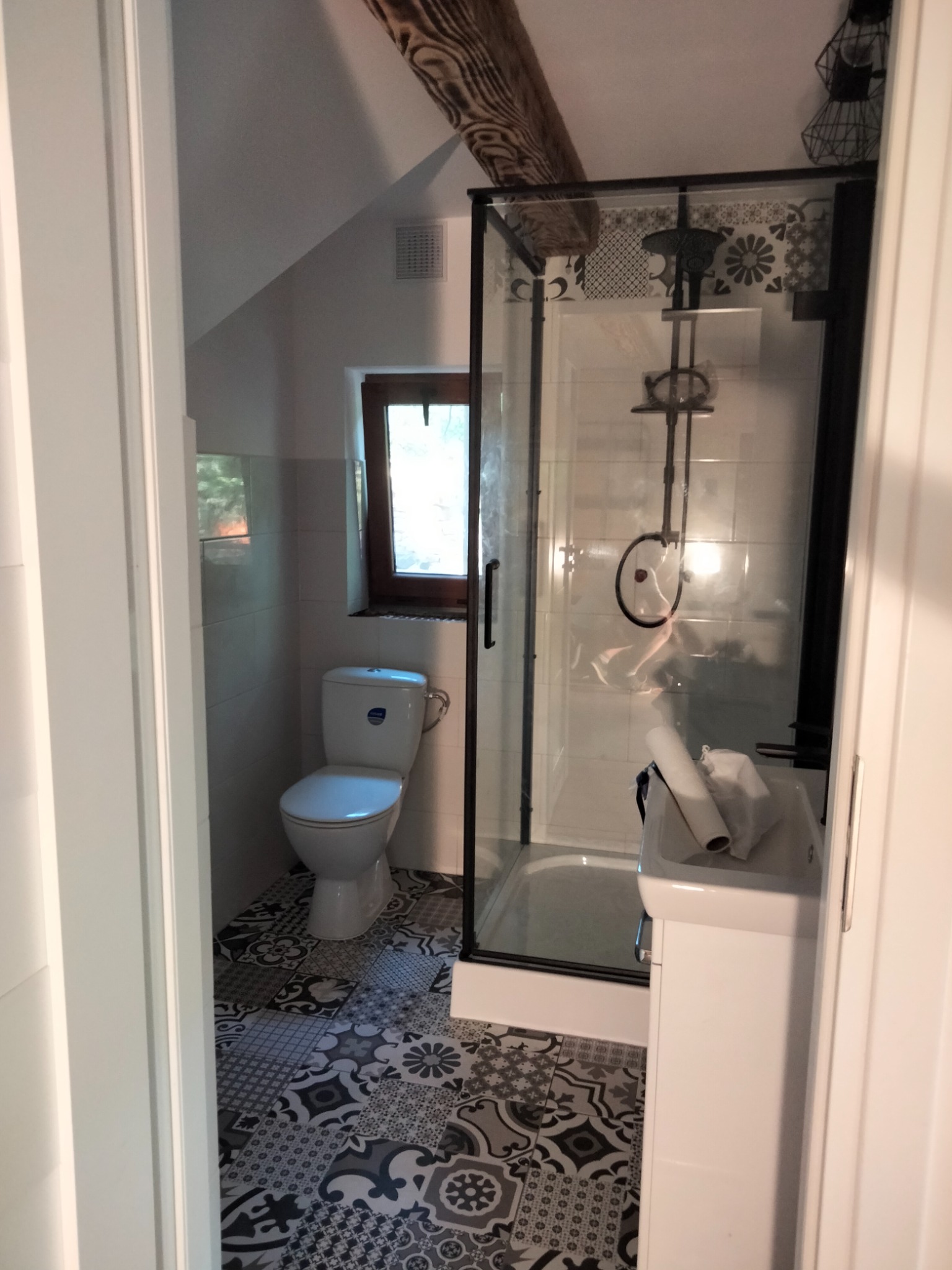 Ukończona łazienka na poddaszu z kabiną prysznicową z czarnymi ramami, toaletą i umywalką na szafce, wykończona mozaikową podłogą i drewnianą belką stropową.