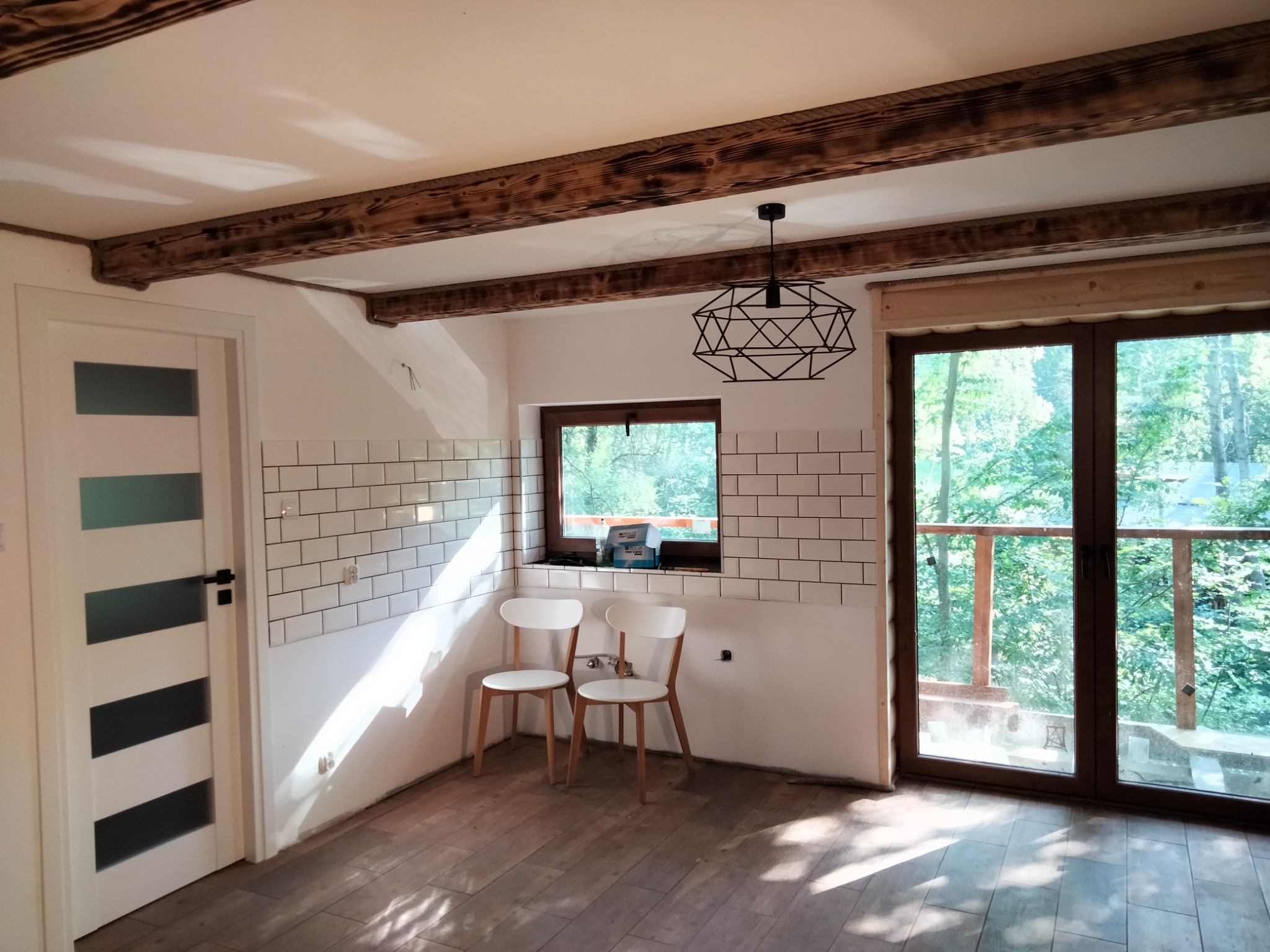 Wnętrze w trakcie remontu: częściowo wyłożona białymi płytkami ściana, drewniane belki stropowe, surowe drewniane wykończenie okna, modernistyczna lampa wisząca, parkiet ułożony we wzór cegiełki...