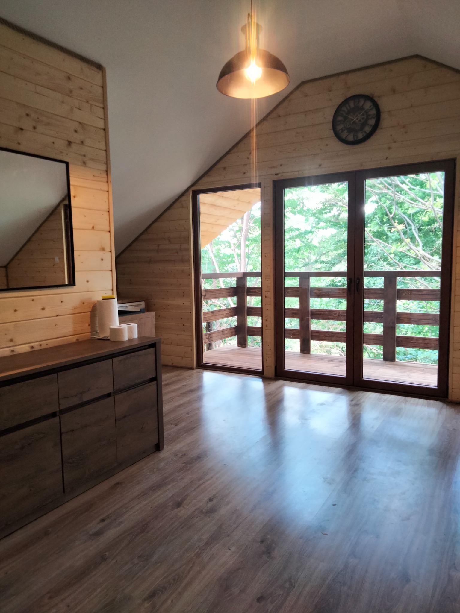 Drewniane wnętrze poddasza z wyjściem na balkon; widoczne panele podłogowe w kolorze drewna, komoda, lustro, zegar na ścianie i lampa wisząca.