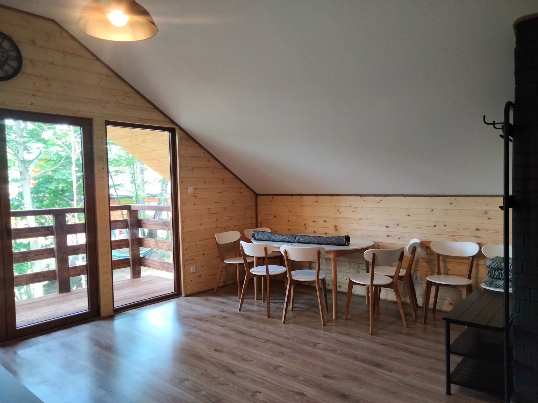 Jasne wnętrze poddasza wykończone drewnem, z dużym oknem balkonowym, okrągłym stołem i krzesłami w stylu skandynawskim oraz panelami podłogowymi imitującymi drewno.