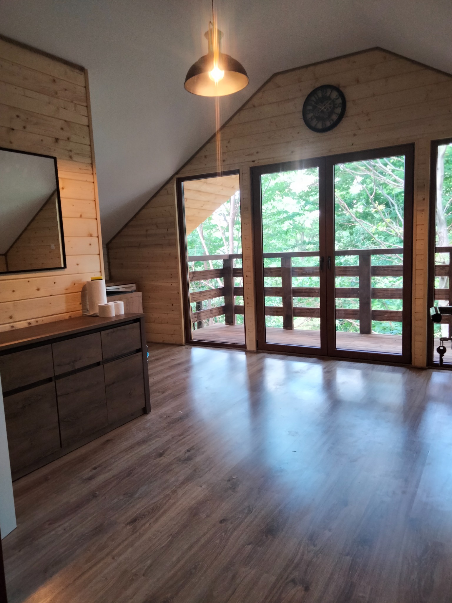 Drewnem wykończone poddasze z panelami podłogowymi, szafką z szufladami i wyjściem na balkon z drewnianą balustradą, widok na zieleń drzew za oknem.
