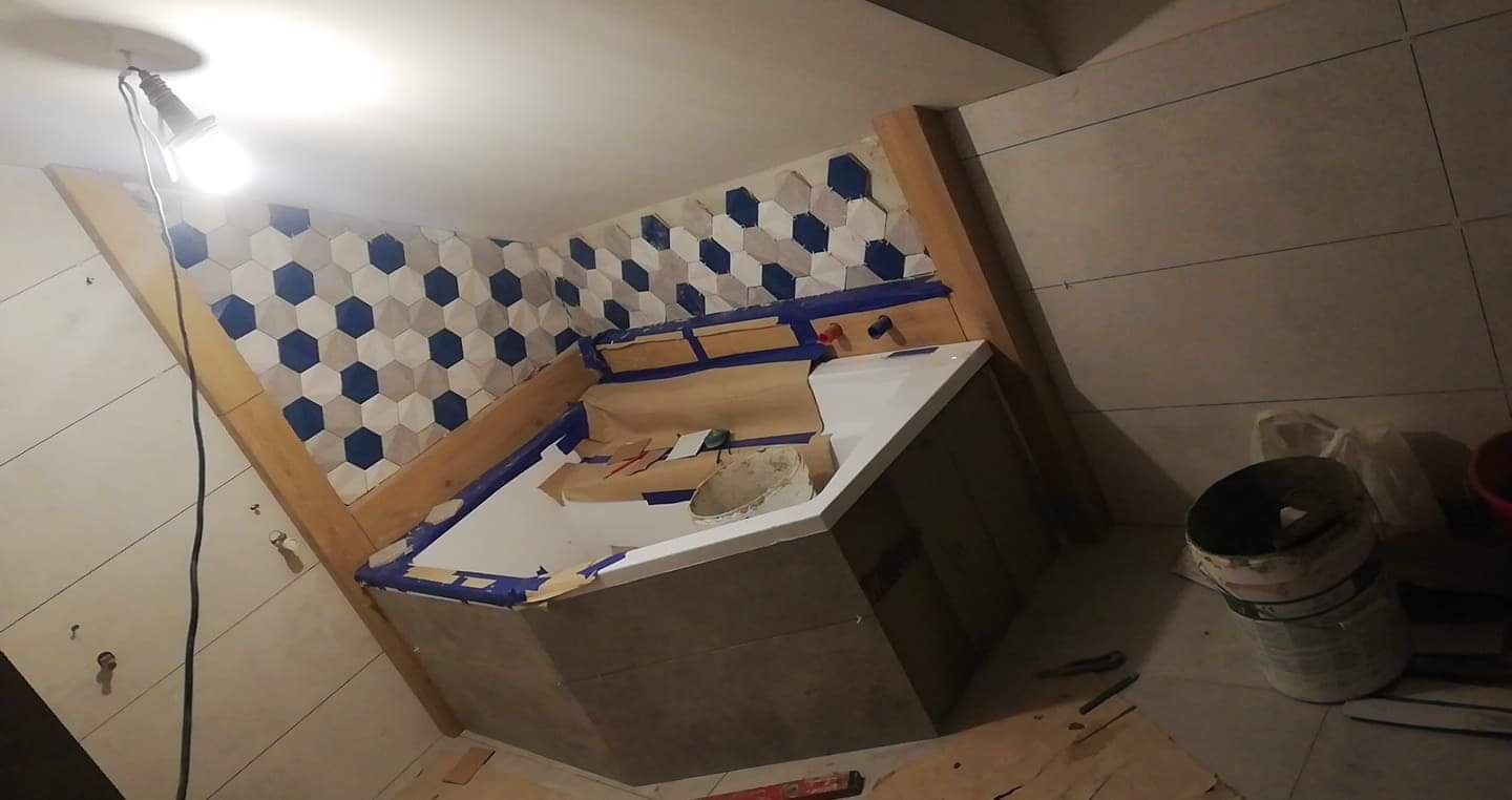 Remont łazienki w trakcie: heksagonalne płytki w odcieniach bieli i niebieskiego na ścianie, wanna zabezpieczona taśmą malarską, widoczna lampa warsztatowa i narzędzia.