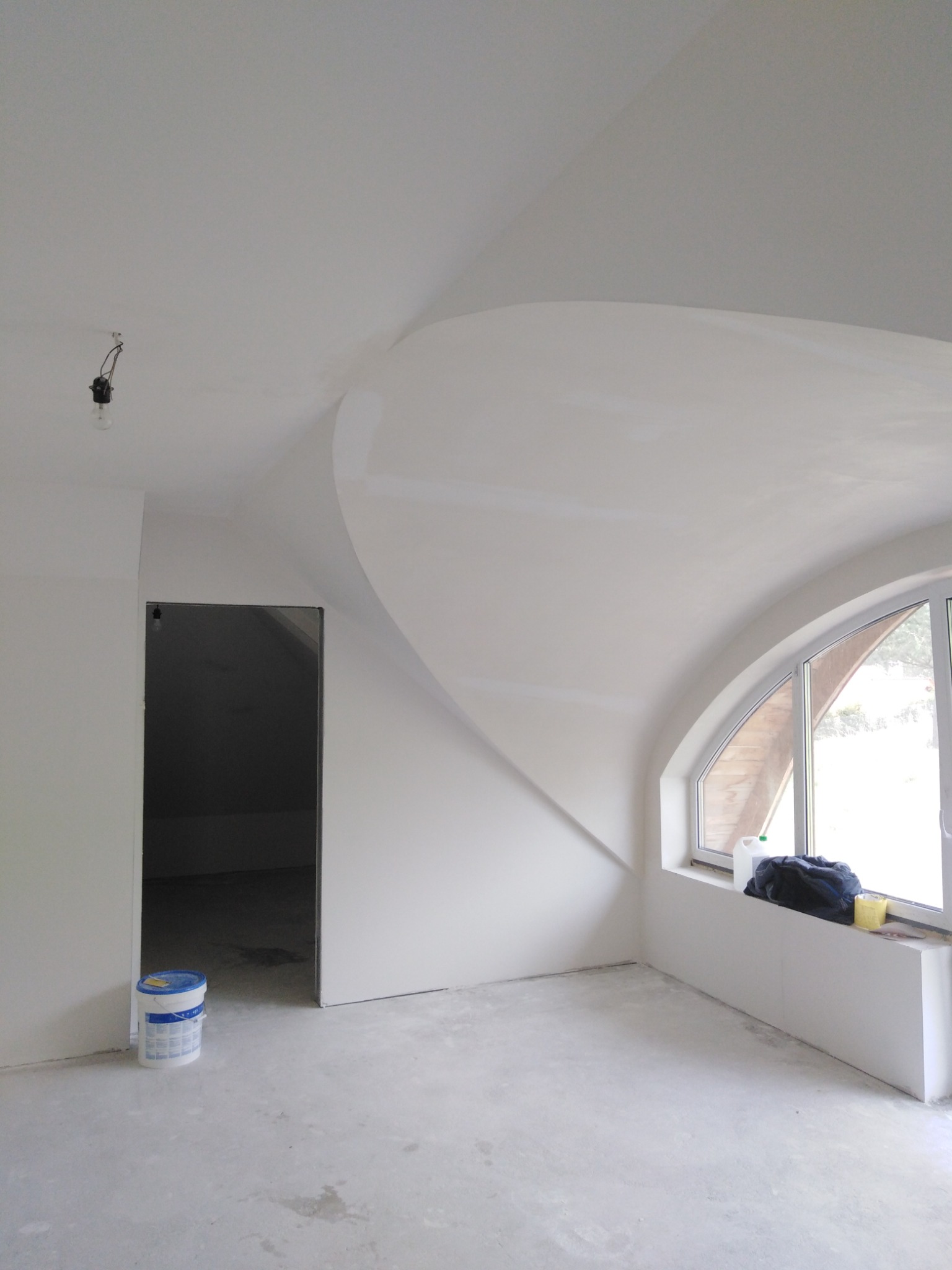 Wnętrze poddasza w trakcie remontu, z charakterystycznym łukowym oknem, widocznym wiadrem z farbą przy wejściu i surowym oświetleniem zwisającym z sufitu.