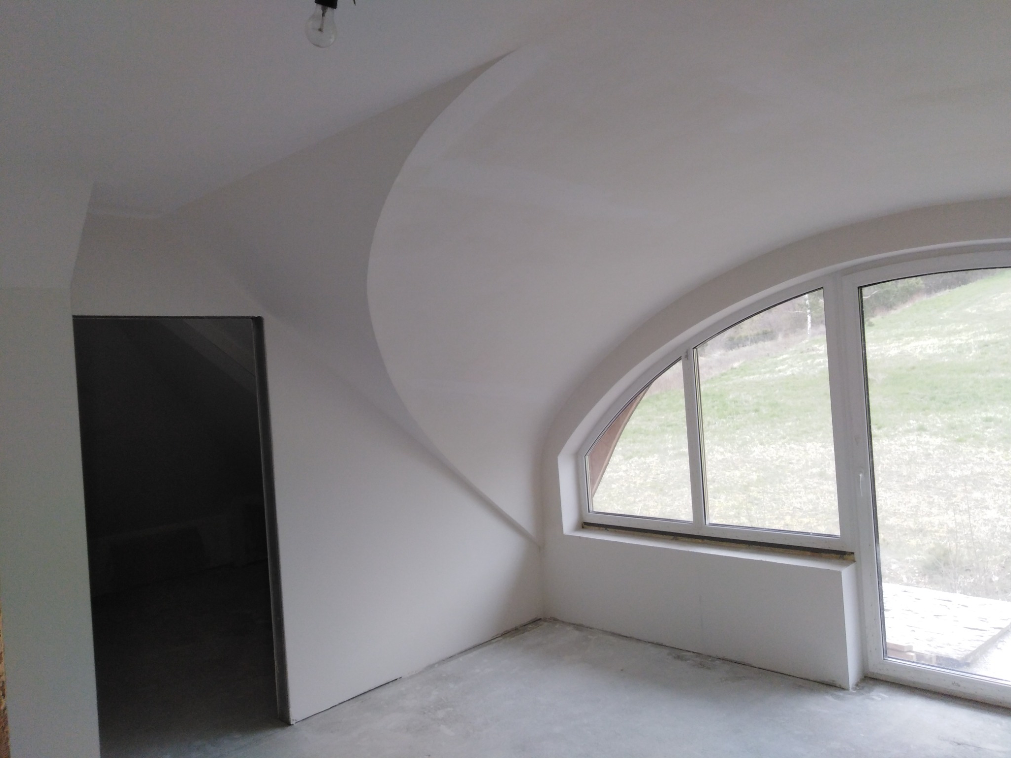 Wnętrze pokoju ze świeżo pomalowanymi ścianami na biało, z nietypowym łukowym przejściem sufitu i oknem z widokiem na zewnątrz, podłoga betonowa.