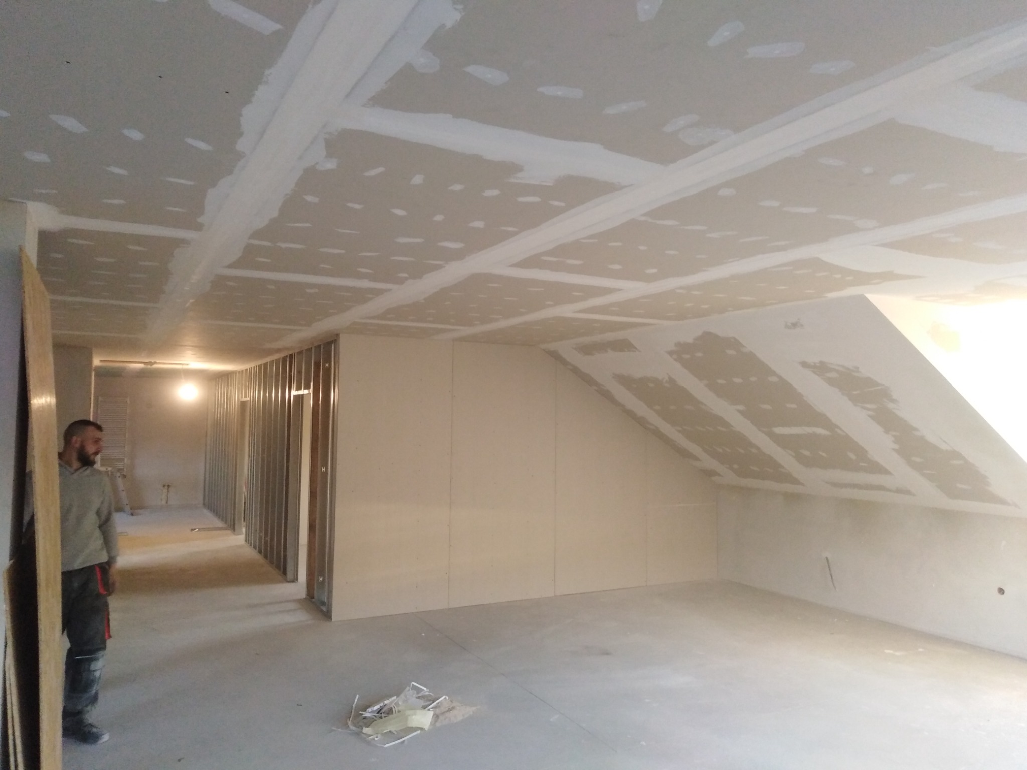 Poddasze w trakcie zabudowy płytami gipsowo-kartonowymi, widoczne szpachlowane łączenia na suficie i ścianach, metalowe profile konstrukcyjne, oraz fragment sylwetki mężczyzny w roboczym ubraniu.