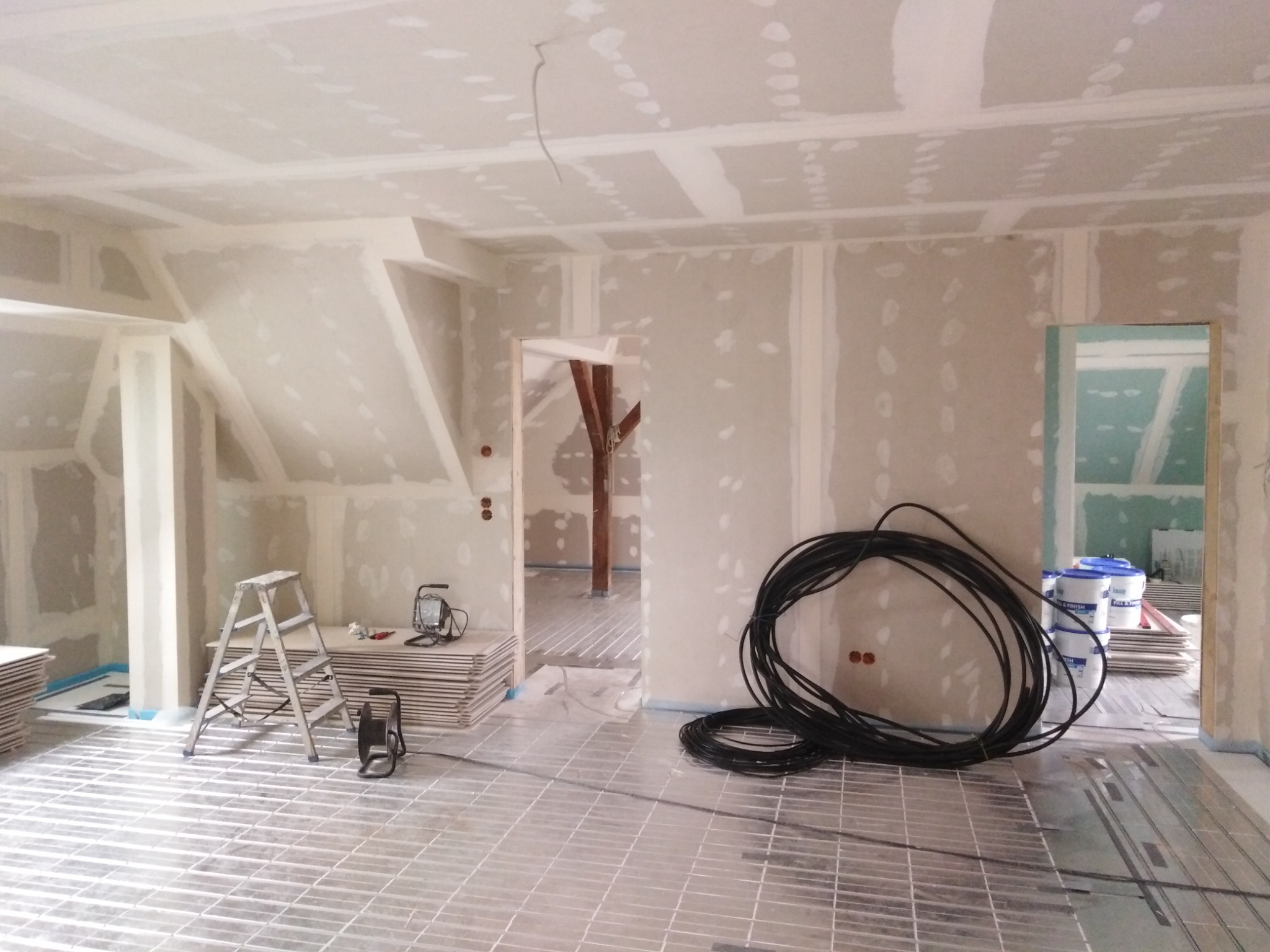 Wnętrze w trakcie remontu: ściany wykończone płytami gipsowo-kartonowymi, podłoga z instalacją ogrzewania podłogowego, drabina malarska, narzędzia i zwój kabli elektrycznych.