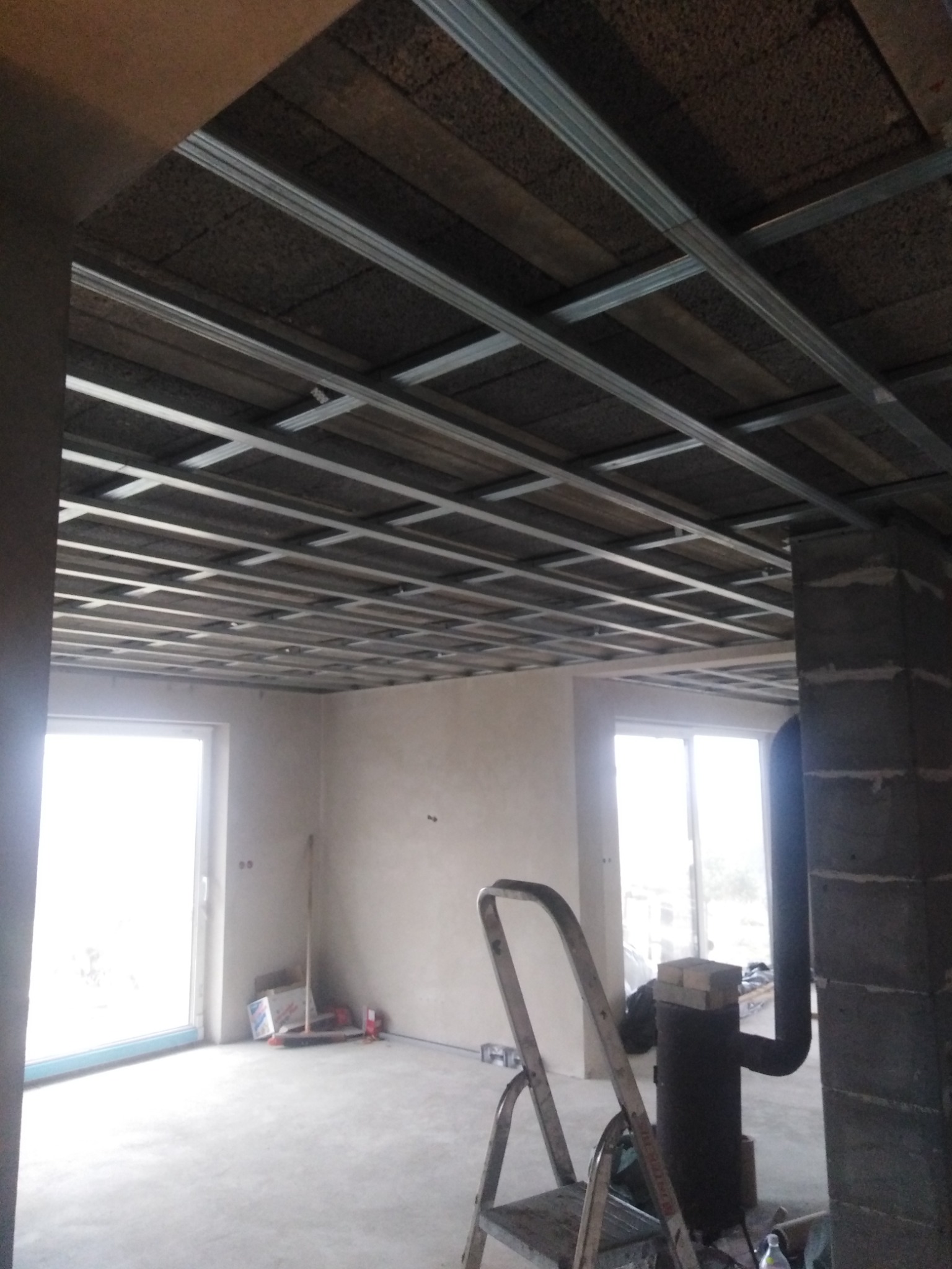 Sufit z metalową konstrukcją i izolacją z wełny mineralnej w trakcie montażu, widoczna drabina, piec typu koza i okno w surowym pomieszczeniu.