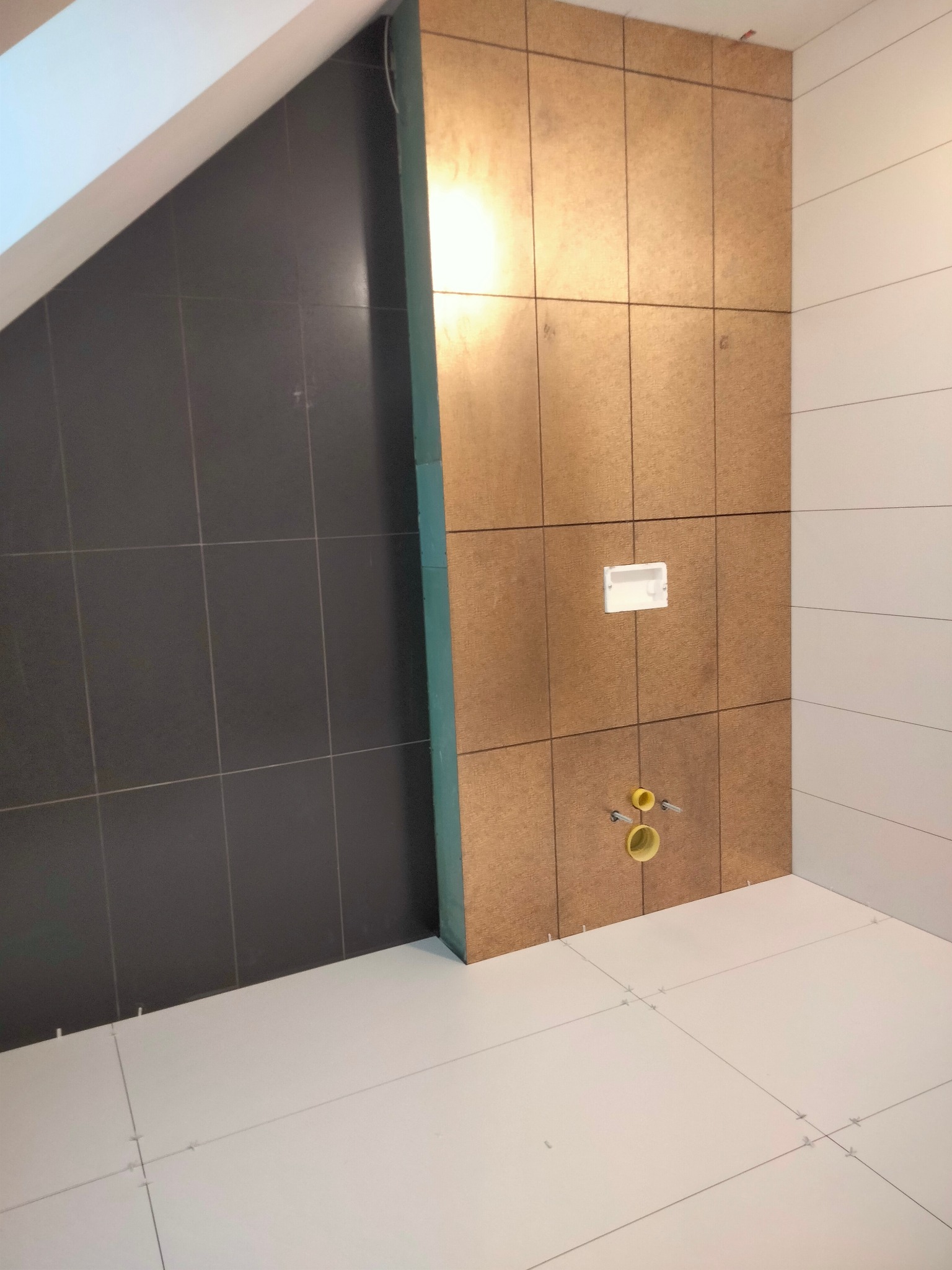 Łazienka w trakcie remontu: ciemnoszare płytki na skośnej ścianie, jasne płytki na podłodze z krzyżykami dystansowymi, płyta OSB na ścianie z instalacją podtynkową WC i przyłączami wodnymi.