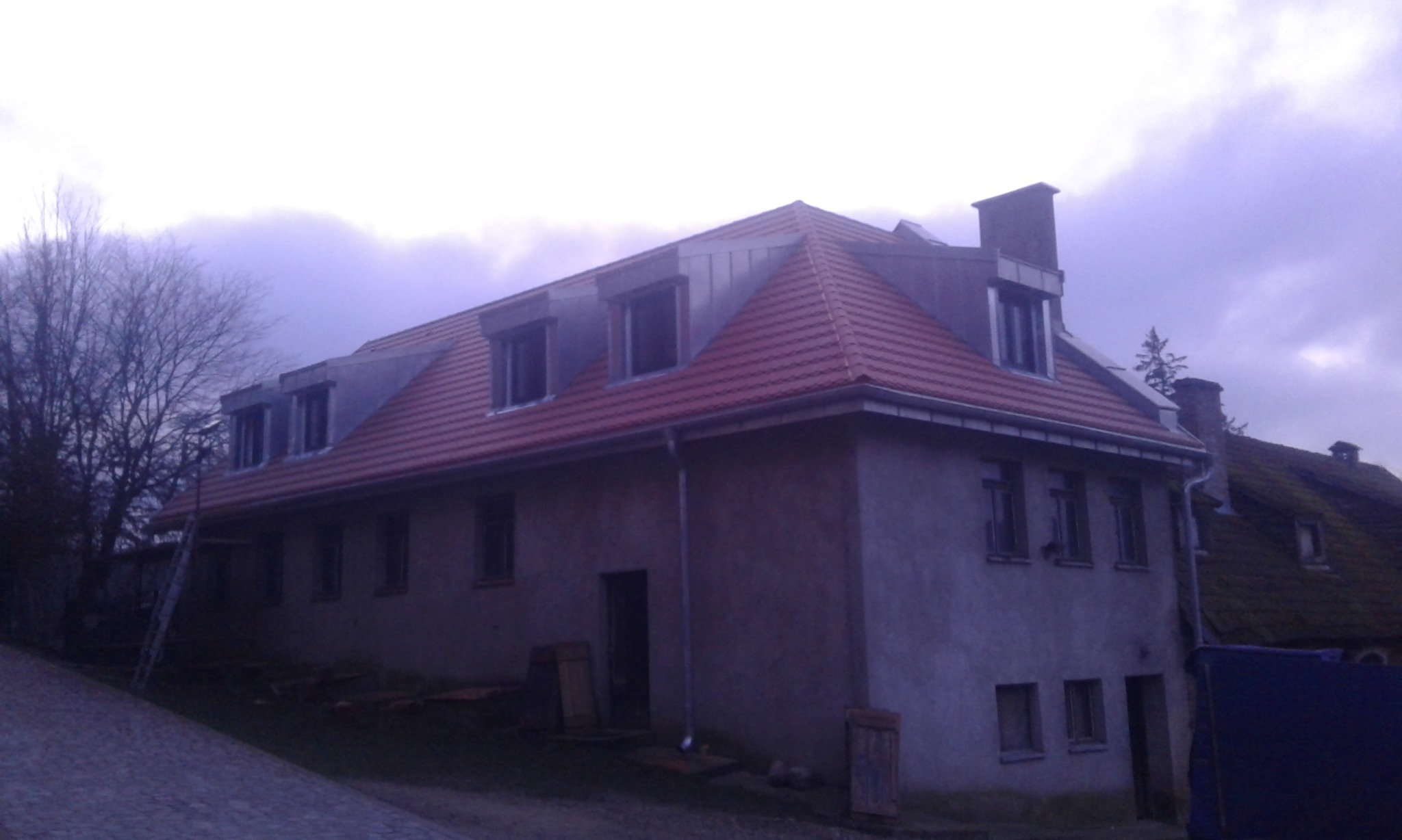 Dwukondygnacyjny budynek z nowym, czerwonym dachem ceramicznym i metalową obróbką blacharską wokół okien dachowych, widoczne rynny, drabina oparta o ścianę, ponure niebo.