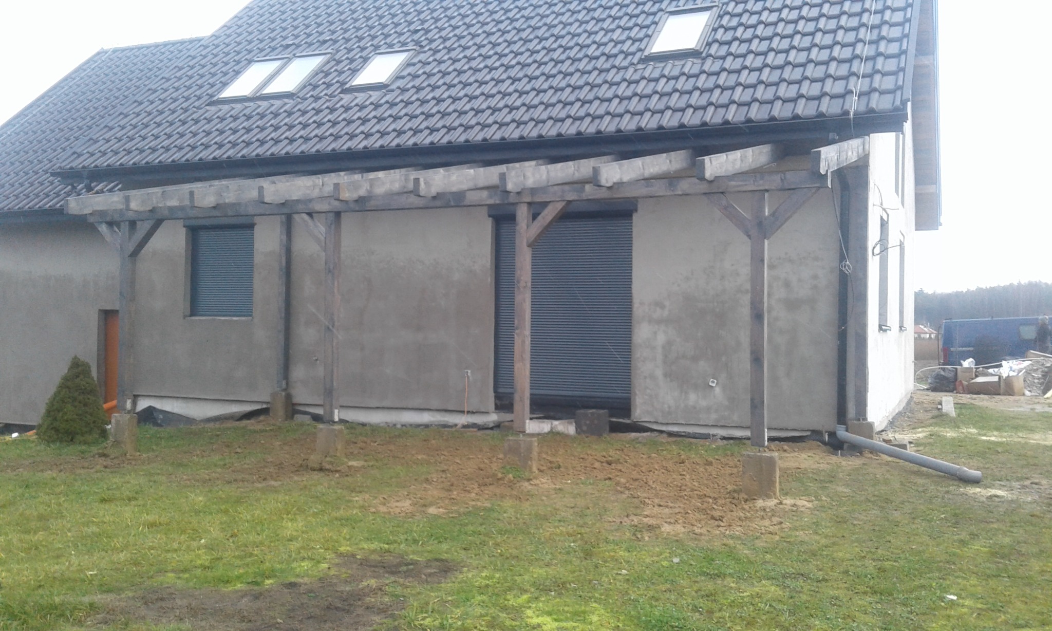 Dom z ciemną dachówką i dobudowanym drewnianym zadaszeniem, widoczne rynny i okna dachowe.