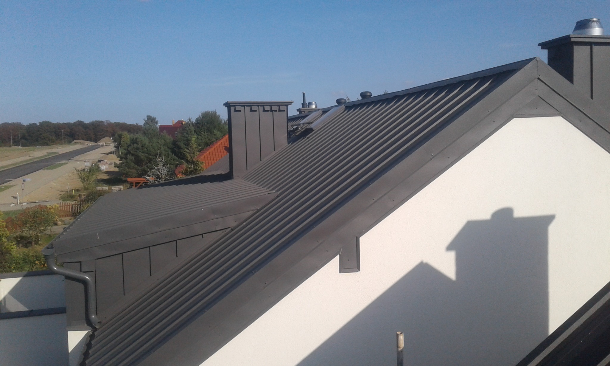 Szary dach z blachy trapezowej z zamontowanym systemem rynnowym, widok z góry na tle błękitnego nieba i zabudowy jednorodzinnej.