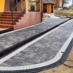 Nowo ułożona, dwutorowa ścieżka z szarej kostki brukowej z białymi i czarnymi obrzeżami prowadząca do schodów wejściowych, czyszczona myjką ciśnieniową, z widocznym stosem kostki i wózkiem...