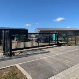 Nowoczesna, przesuwna brama wjazdowa z czarnych, pionowych profili na posesji z budynkami gospodarczymi i sprzętem budowlanym w tle, widoczna kostka brukowa podjazdu.