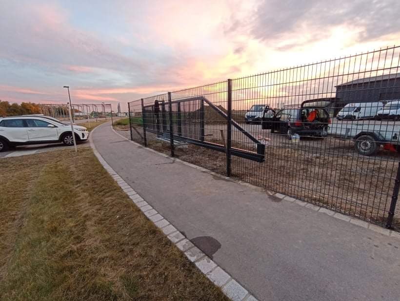 Nowo zainstalowane, czarne, metalowe ogrodzenie panelowe z przesuwną bramą wjazdową, widoczne z boku ścieżki, na tle zachodzącego słońca i zaparkowanych pojazdów.