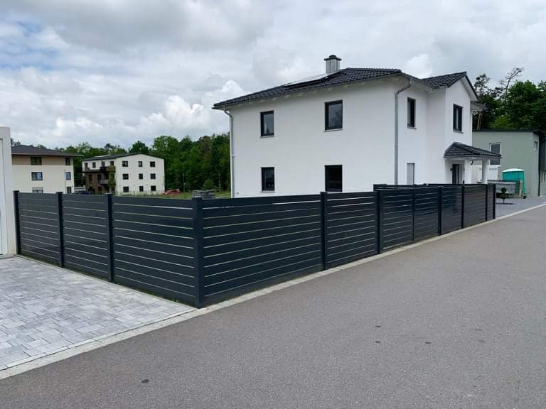 Nowoczesne, poziome ogrodzenie panelowe w kolorze antracytowym, otaczające posesję z białym domem jednorodzinnym i szarą kostką brukową na podjeździe.