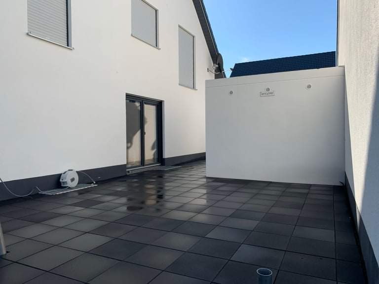 Świeżo ułożona, mokra, grafitowa kostka brukowa na tarasie przylegającym do budynku z białymi ścianami i czarną stolarką okienną. Widoczny zwinięty wąż do wody oraz fragment instalacji elektrycznej...