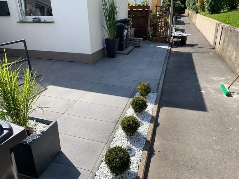 Szara kostka brukowa ułożona na posesji, obok chodnika z zieloną szczotką, dekoracyjne kuliste krzewy oddzielone białymi kamieniami.