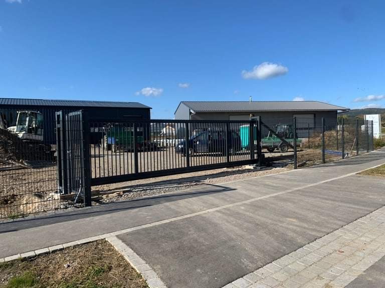 Nowoczesna, przesuwna brama wjazdowa z czarnych, pionowych profili na posesji z budynkami gospodarczymi i sprzętem budowlanym w tle, widoczna kostka brukowa podjazdu.